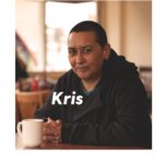 A photo of Kris, a brown skinned person with close cropped dark hair, sat at a table with a white coffee cup and a dark hoody, softly smiling to the camera with their name Kris printed across the picture