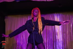 The performer Shakona Fire in standing on a stage with arms outstretched to each side, in front of a stage curtain.  Shakona is wearing fish net tight and a navy blue jacket with a belt. Shakona has long braids which fade from dark to light orange, and septum piercing. Tha image has a purple hue from stage lighting. The bottom left corner contains the watermark Wotever World, Henri T, (@henri t art)