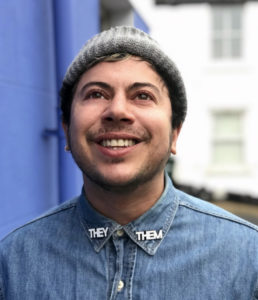 Fox Fisher is looking upwards towards the sky and is standing in front of a blue and a white building. Fox is wearing a grey beany hat and a blue shirt that has the words they and them one each collar. Fox is smiling and has stubble.