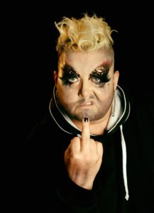 The image contains Ingo Cando with messy blonde hair on top and shaved sides. Ingo is wearing glittering eye make up and big black eyelashes. Ingo has facial hair shadow applied with make up. Ingo's expression is snarling. Ingo is wearing a black hoody and is holding up a fist with the middle finger extended. The finger nail of the middle finger is painted black. 