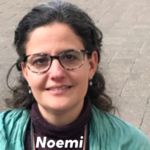 A photo of Noemi with shoulder length hair, glasses a, green shirt and earrings, softly smiling to the camera and with their name Noemi printed at the bottom