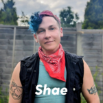 a portrait photo of Shae, a white person with blue and red hair wearing a red bandana and black and green tops. Their name Shae is printed at the bottom